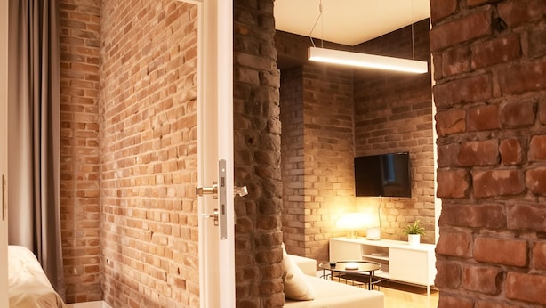A modern room with exposed brick walls, a flat-screen TV mounted on the wall, a white sofa, and a small round table. - Cracow Fancy Brick #4 - Old Town (Kraków)