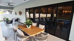 A patio area with a wooden table and wicker chairs, a potted plant, and a glass door leading to a pool area. - New spacious luxury 4-bedroom villa in gated development close to beaches. (Canaan)