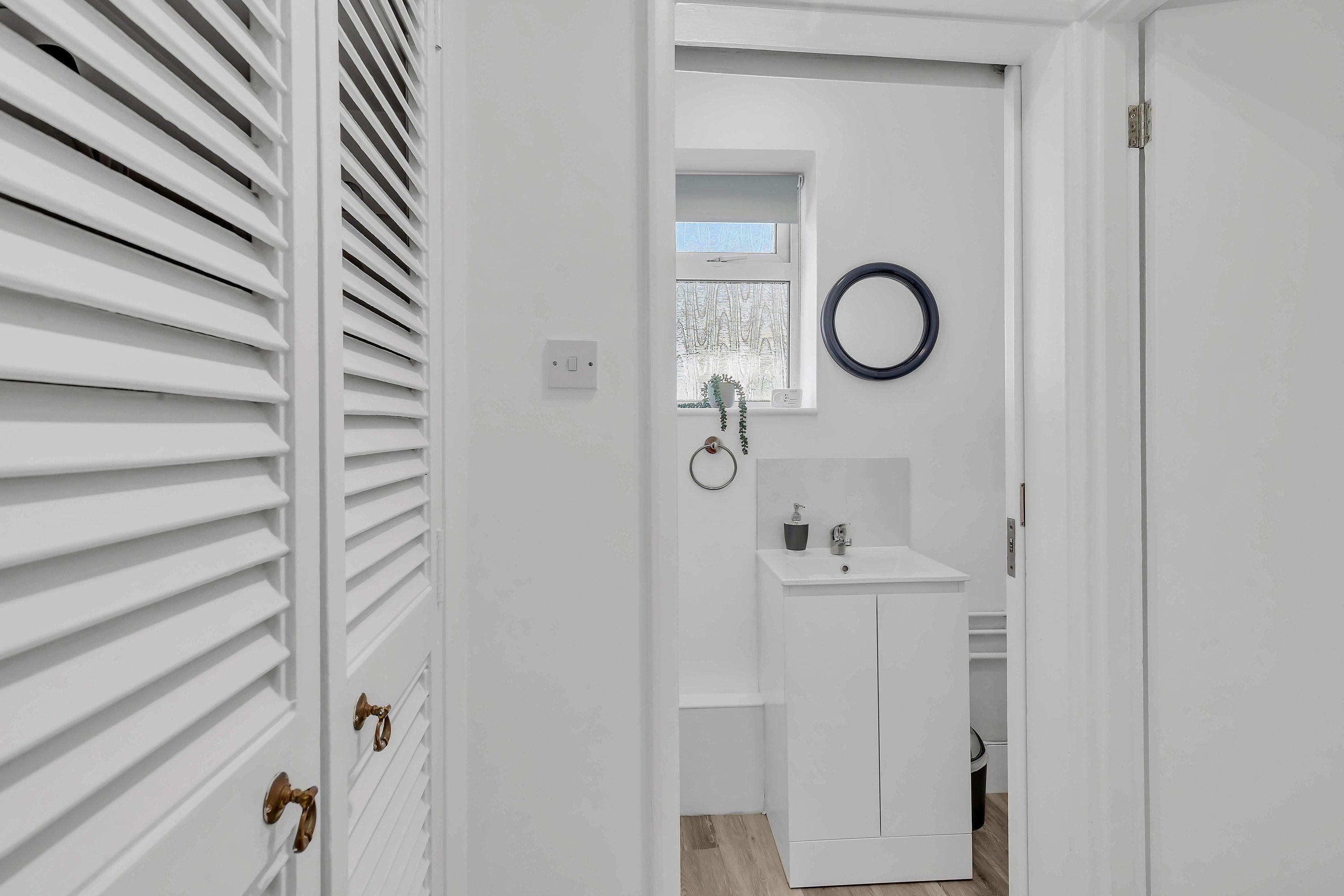 A narrow hallway with white walls, a white door with brass handles, and a window with blinds.