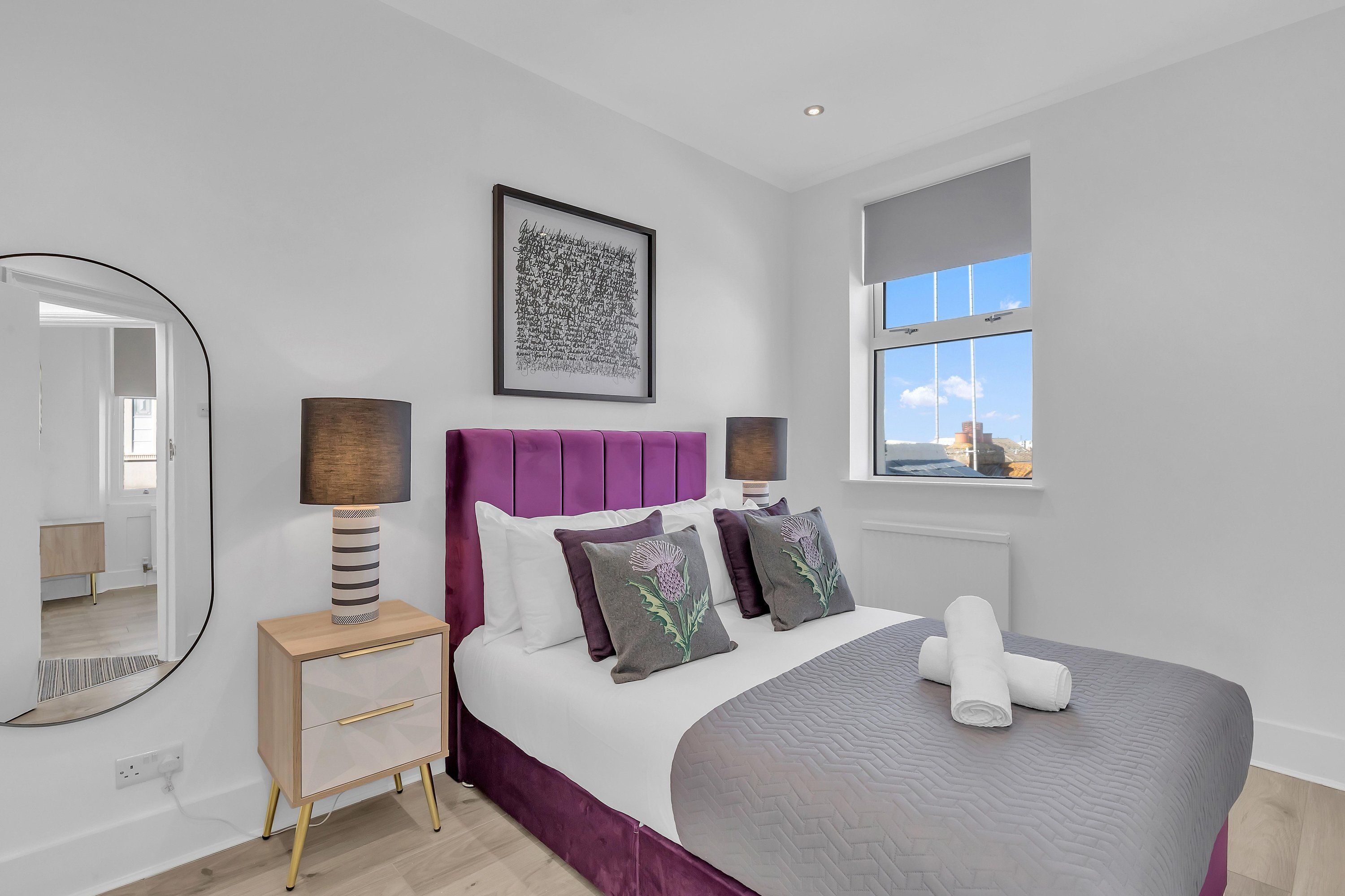 A modern bedroom with a large bed, a bedside table, a mirror, and a window with a view.