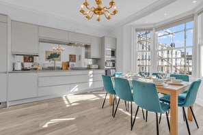 A modern kitchen with white cabinetry, a central island, and a dining area with teal chairs and a wooden table. - City Chic Townhouse By My Getaways (Brighton)