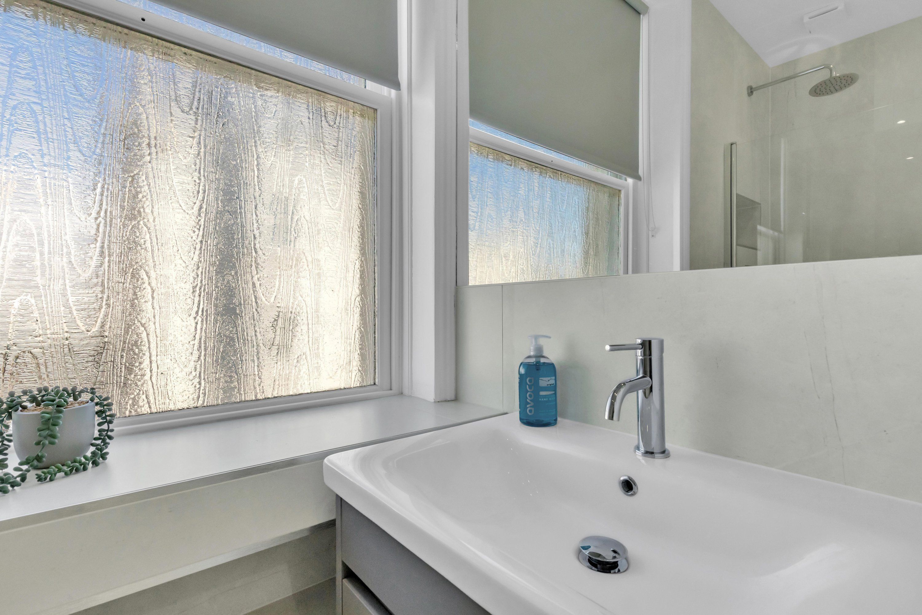 A modern bathroom with a large window, a white sink, a mirror, and a shower area.