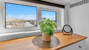 Un bureau en bois, sur lequel se trouvent un pot de fleurs en osier, un objet circulaire, et une vue sur un port de plaisance, visible à travers deux fenêtres.