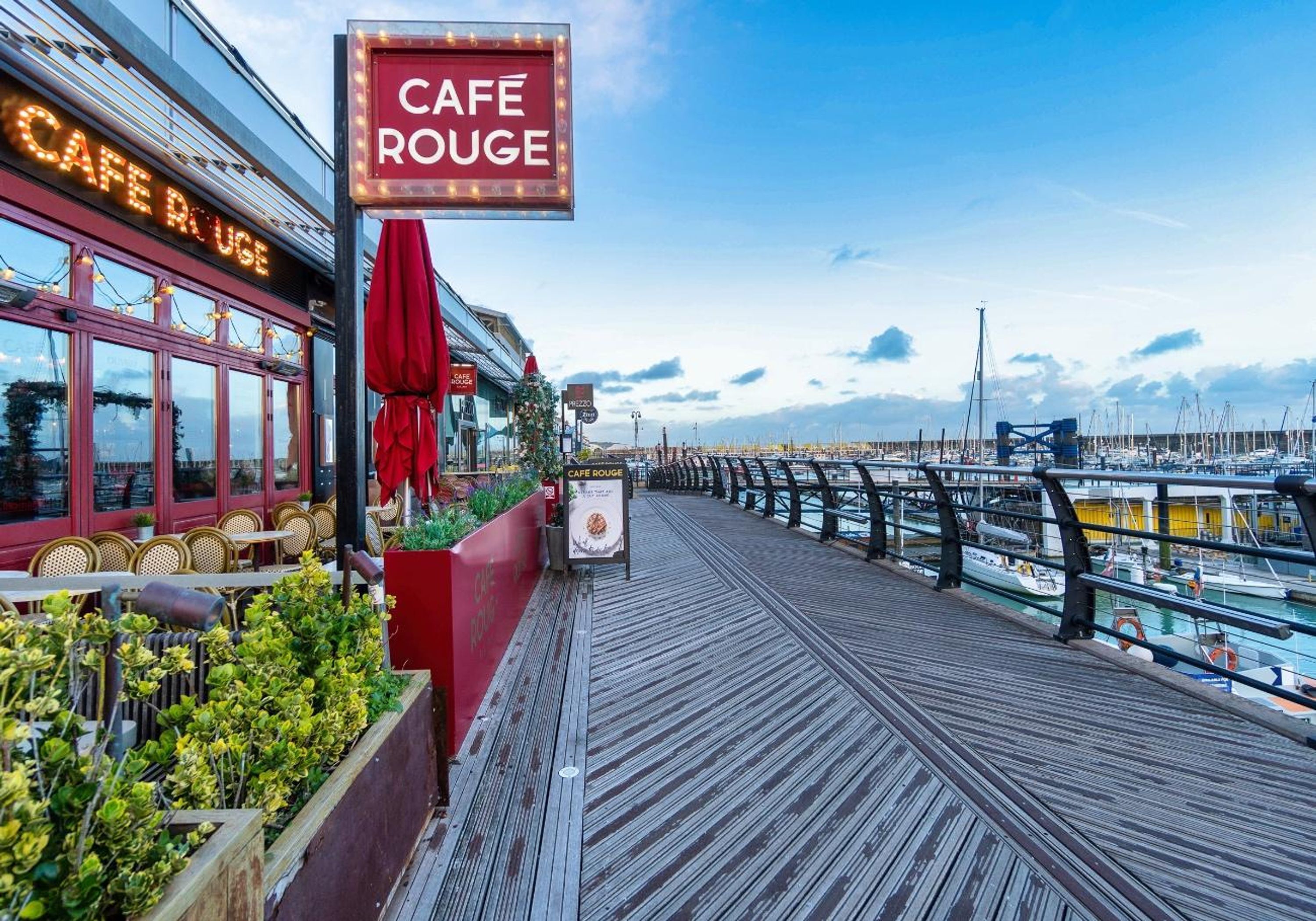A wooden deck with a cafe named "Cafe Rouge" and a view of boats in the background.