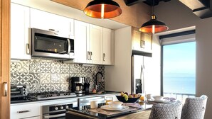 A modern kitchen with white cabinets, a black stove, and a microwave. There is a dining area with patterned chairs and a view of the sea through a large window.