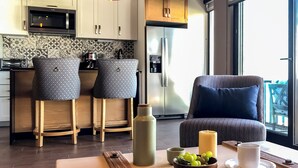 A modern kitchen with a microwave, refrigerator, and two patterned chairs. A large window provides natural light, and a table with a candle and a bowl of grapes is visible.
