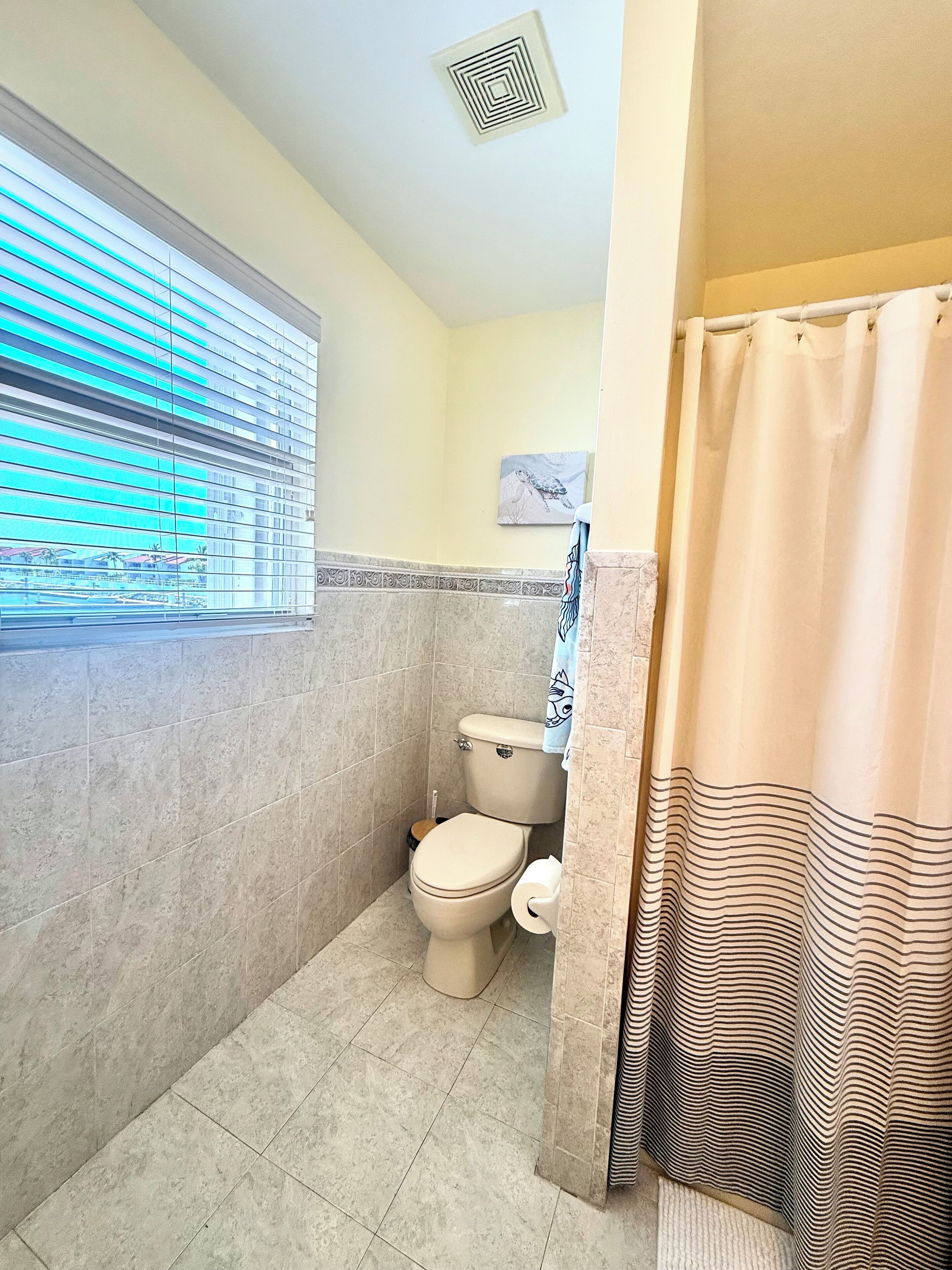 Une salle de bain équipée de toilettes, d’une fenêtre avec stores, d’un rideau de douche et d’une bouche d’aération fixée au mur.