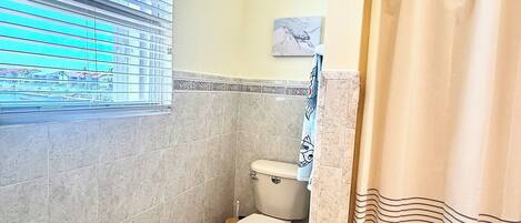 Une salle de bain équipée de toilettes, d’une fenêtre avec stores, d’un rideau de douche et d’une bouche d’aération fixée au mur.
