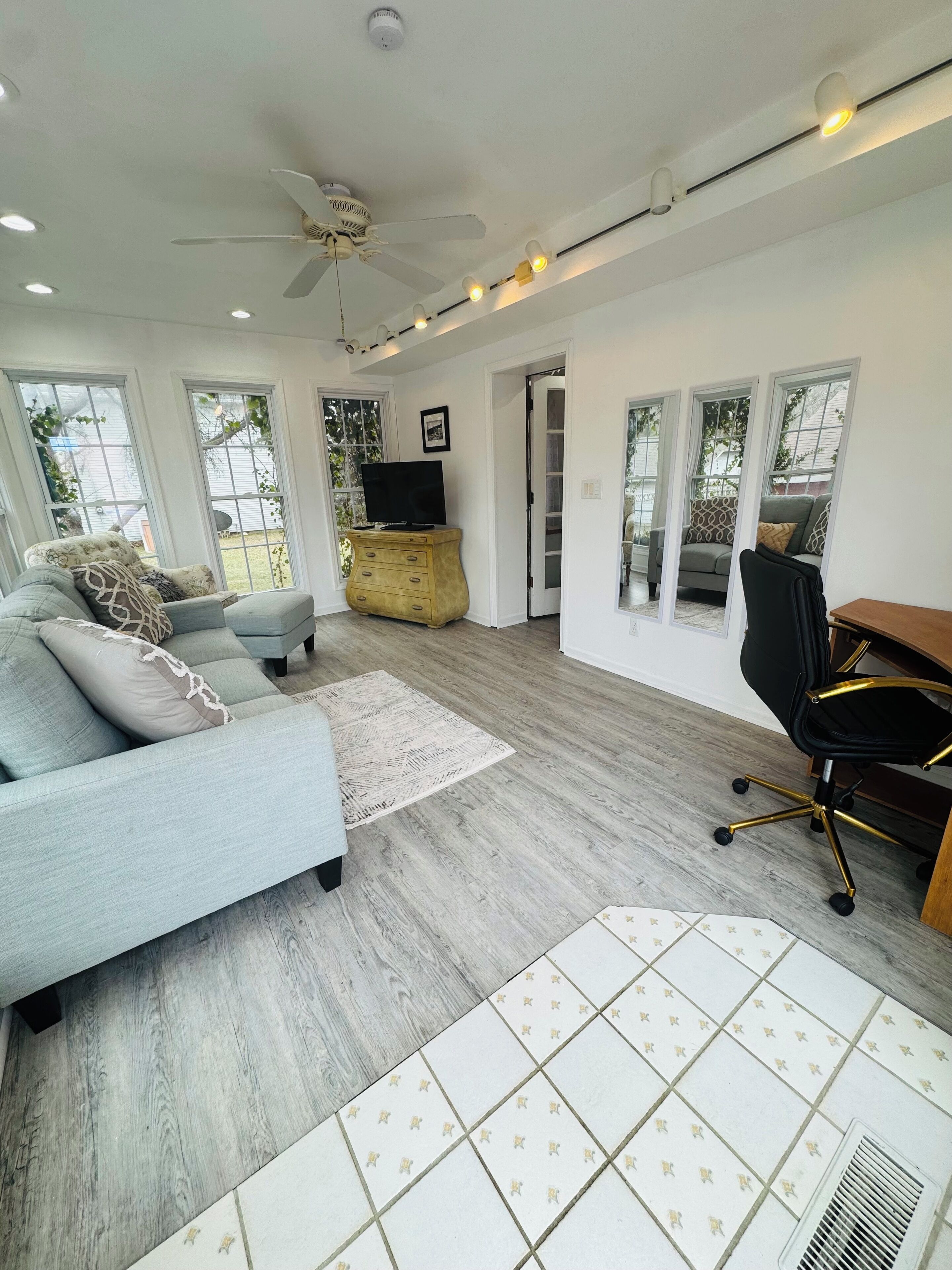 A spacious living room with a light blue sofa, a wooden cabinet, a black office chair, and a ceiling fan.
