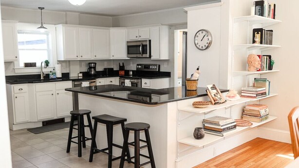 A modern kitchen with white cabinets, a central island with stools, a microwave, and a clock on the wall. - Dutch Retreat • Near Lakes • Walk Town (Bridgton)