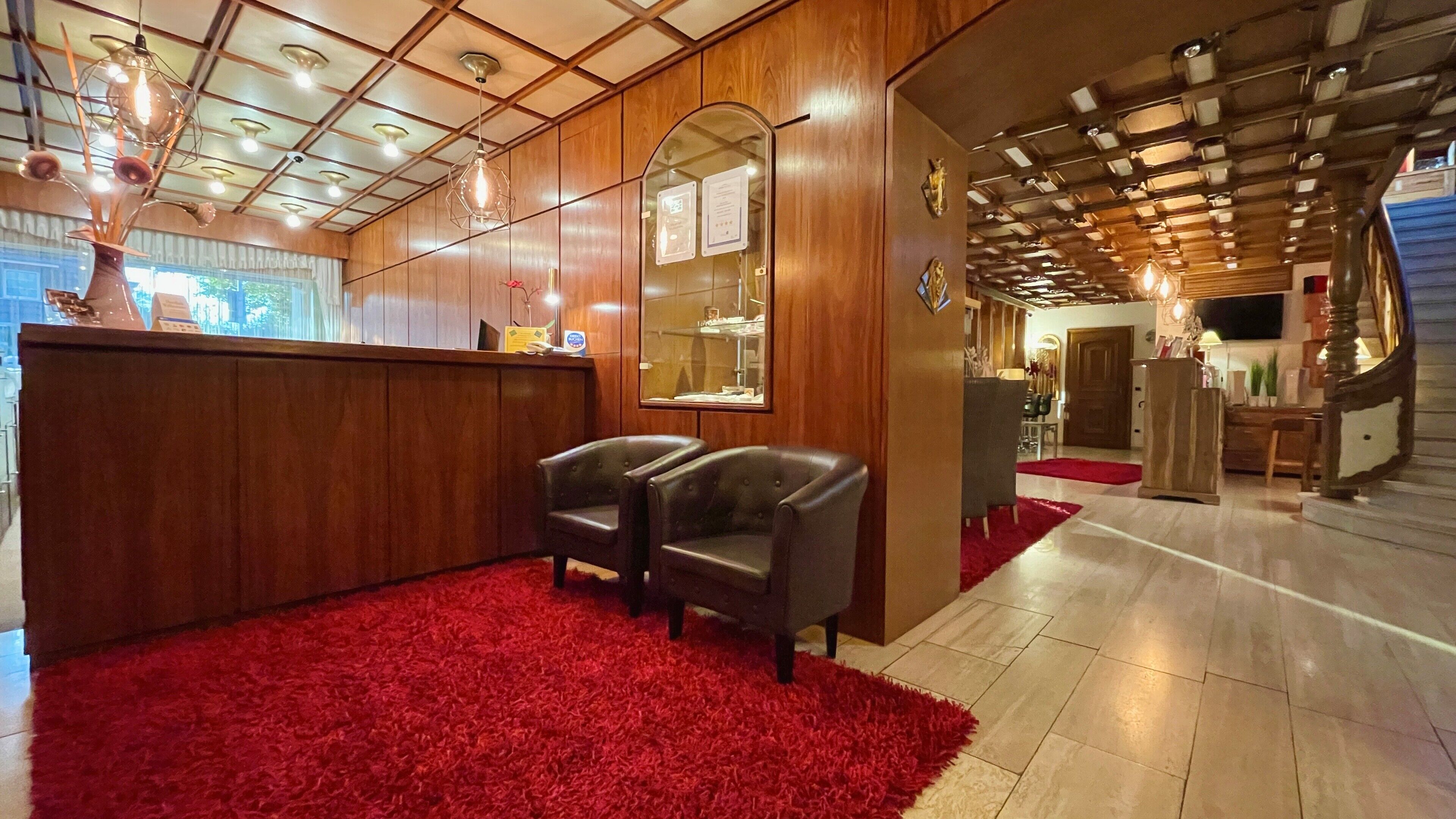Un espace d’accueil avec un bureau en bois, deux fauteuils en cuir, une moquette rouge et un plafond avec des poutres apparentes et des luminaires.