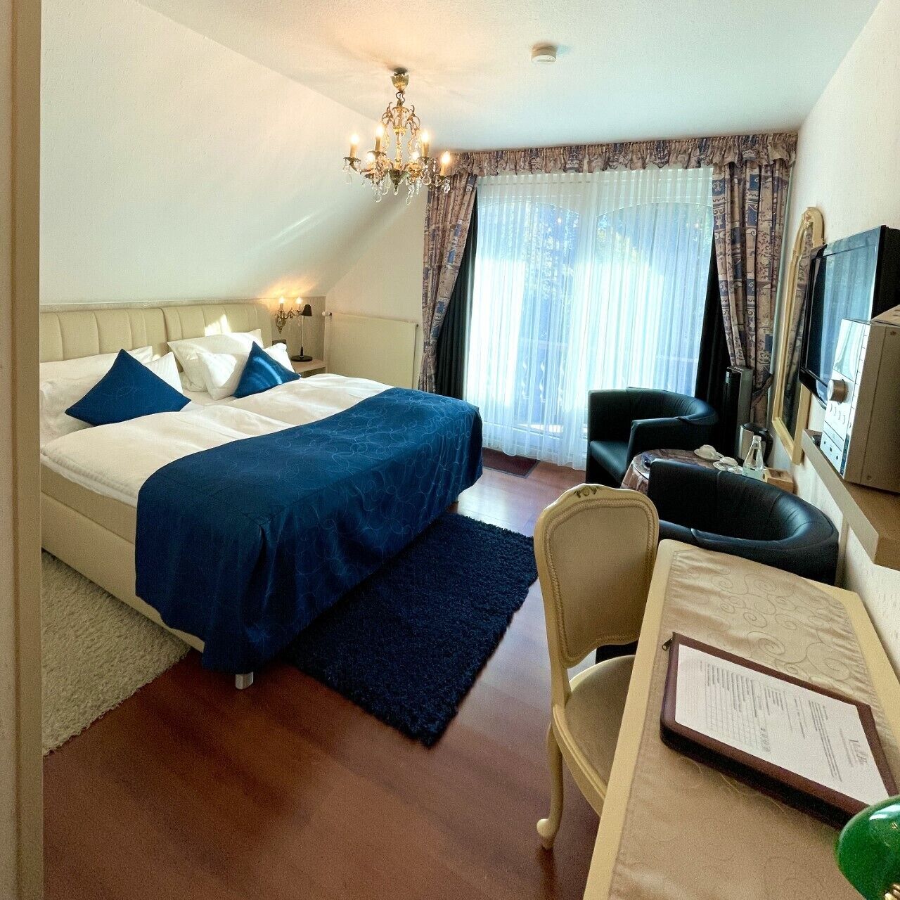 A hotel room with a double bed, a desk with a chair, a television, and a chandelier.