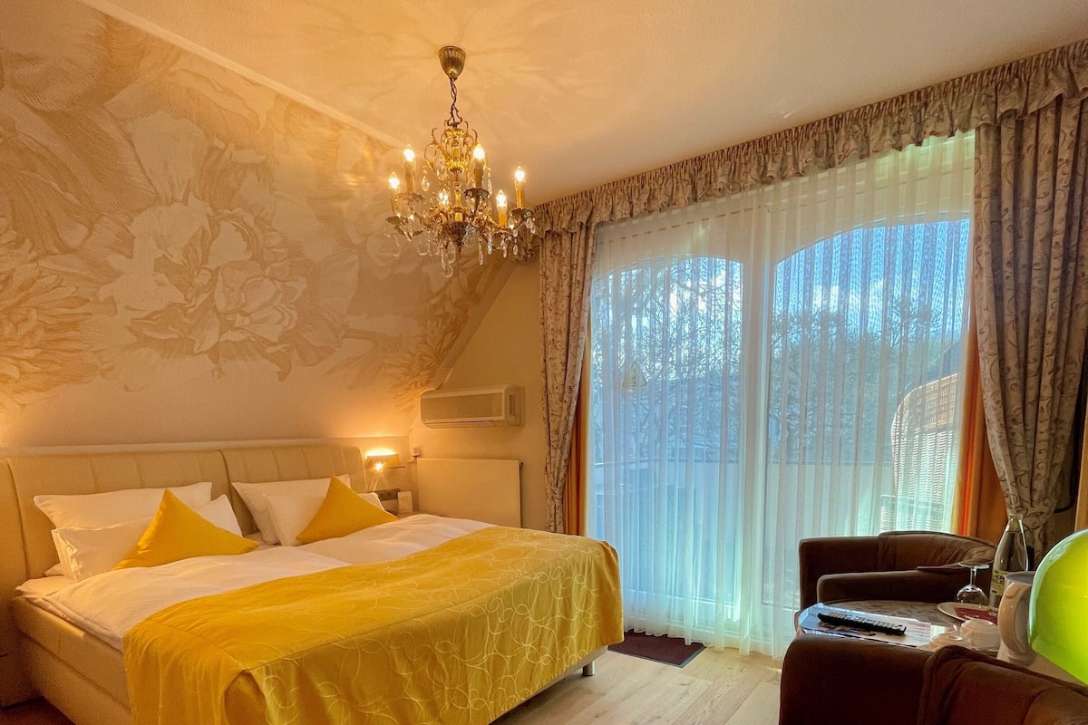 A hotel room with a large bed, a desk, and a chair. There is a chandelier above the bed and a window with curtains.