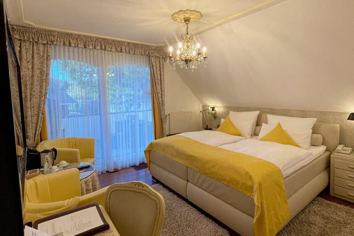 A hotel room with a large bed, a desk with a chair, a chandelier, and a view of the outdoors through the window.