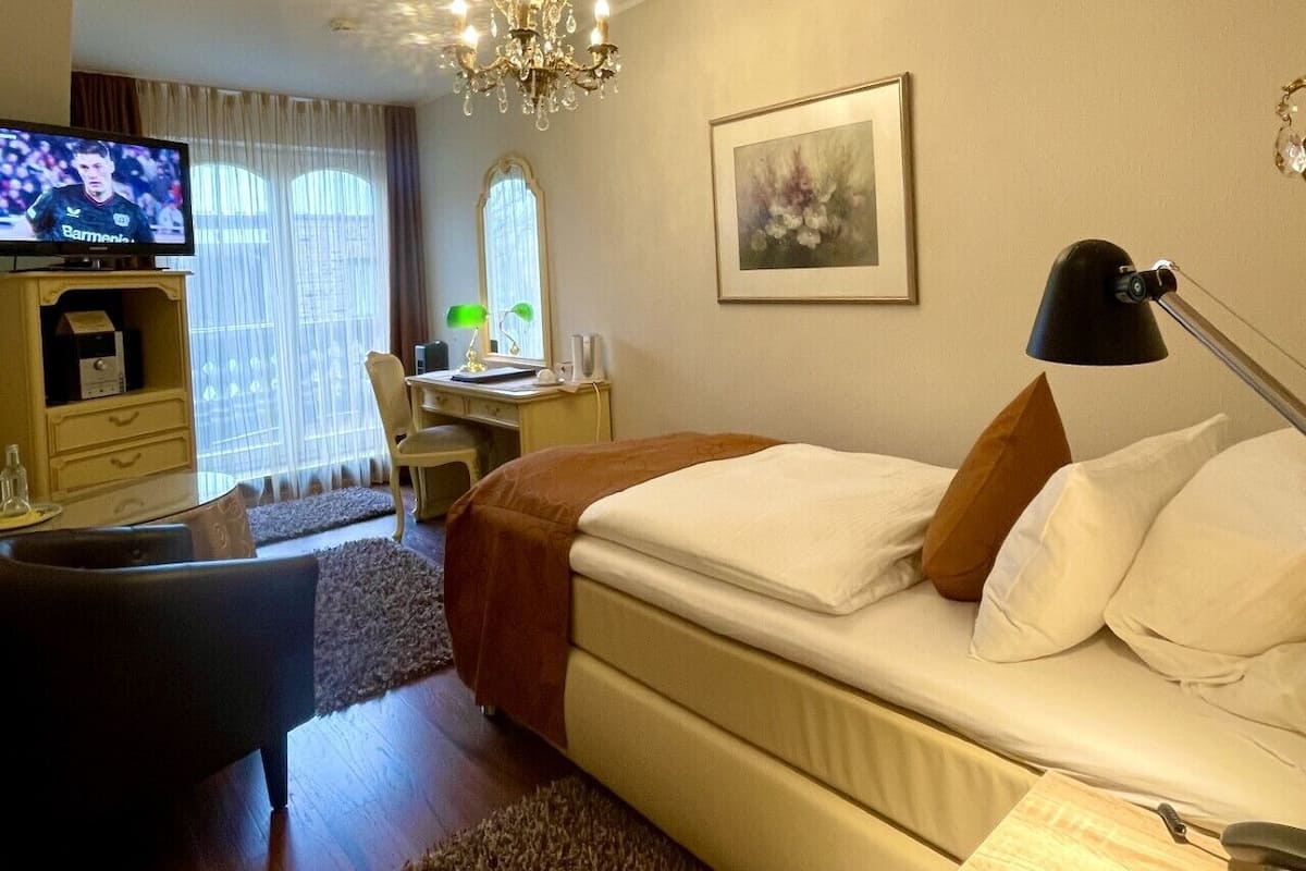 A hotel room with a bed, a desk with a chair, a television, and a chandelier.