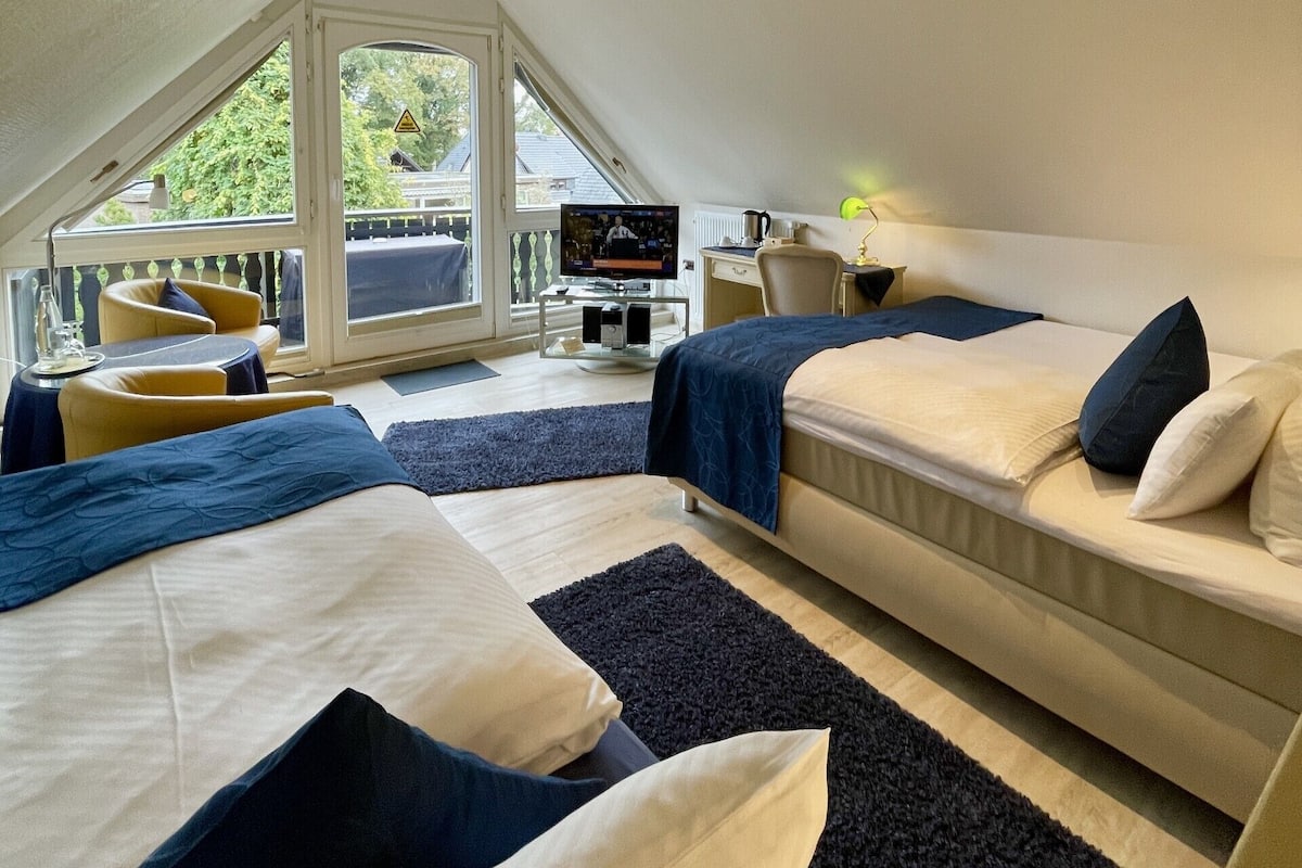 A modern hotel room with two beds, a desk, a television, and a balcony with a view of greenery.