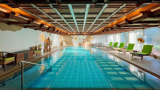 A large indoor swimming pool with a tiled floor, surrounded by lounge chairs and a wooden ceiling structure. - Villa am Park (Bad Zwischenahn)