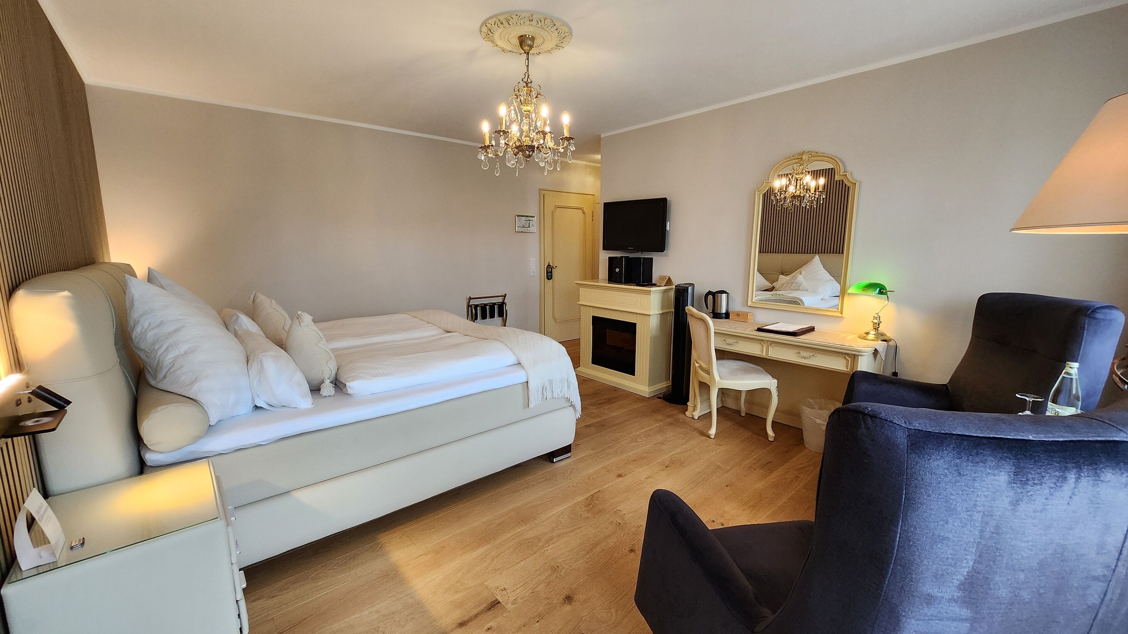 A hotel room with a bed, a desk, a chair, a television, a mirror, and a chandelier.