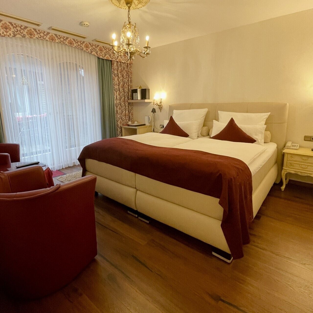 A hotel room with a large bed, a chandelier, a desk with a lamp, and a view of the outside through the window.