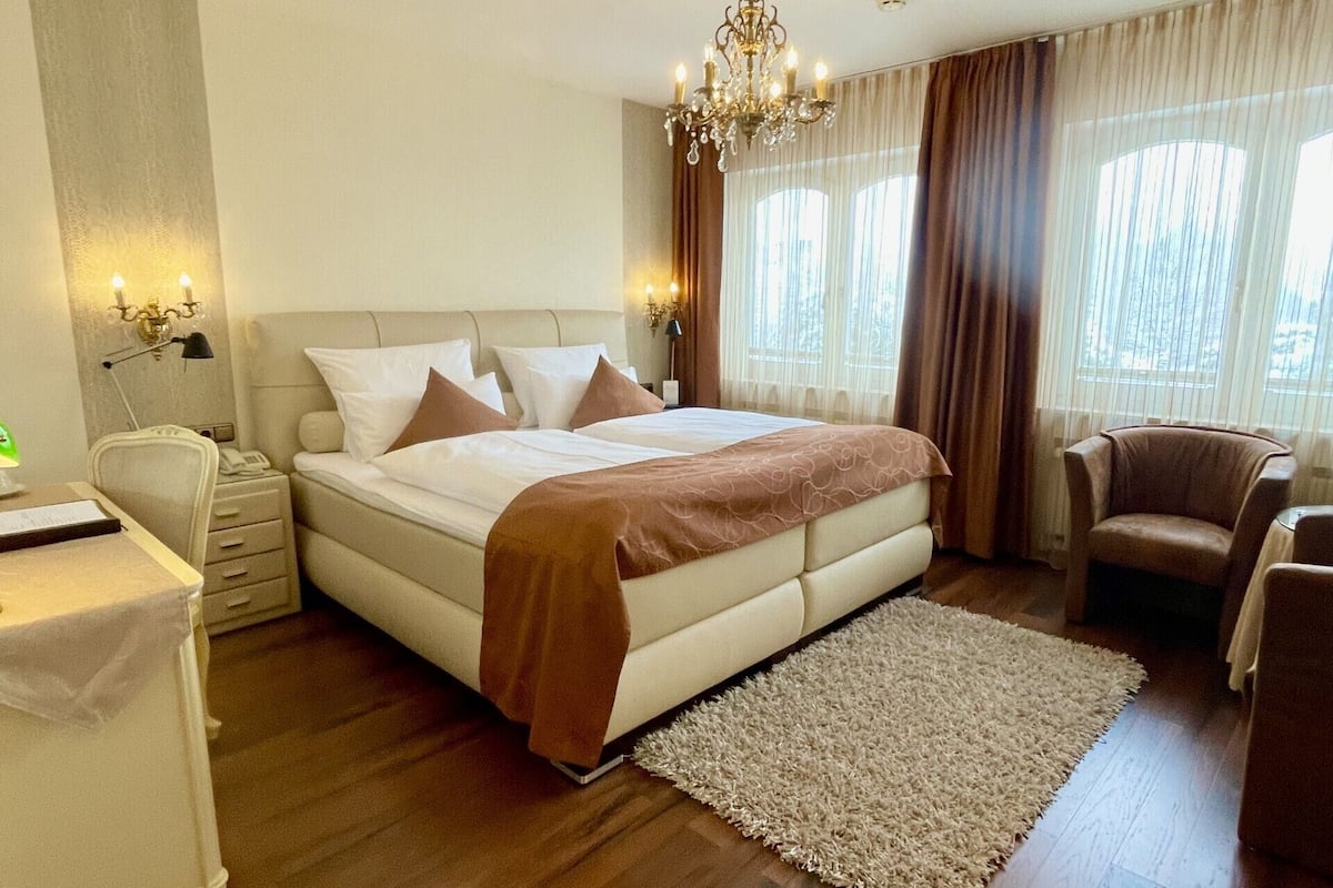 A bedroom with a large bed, a desk, a chair, a chandelier, and a view of the outdoors through the window.
