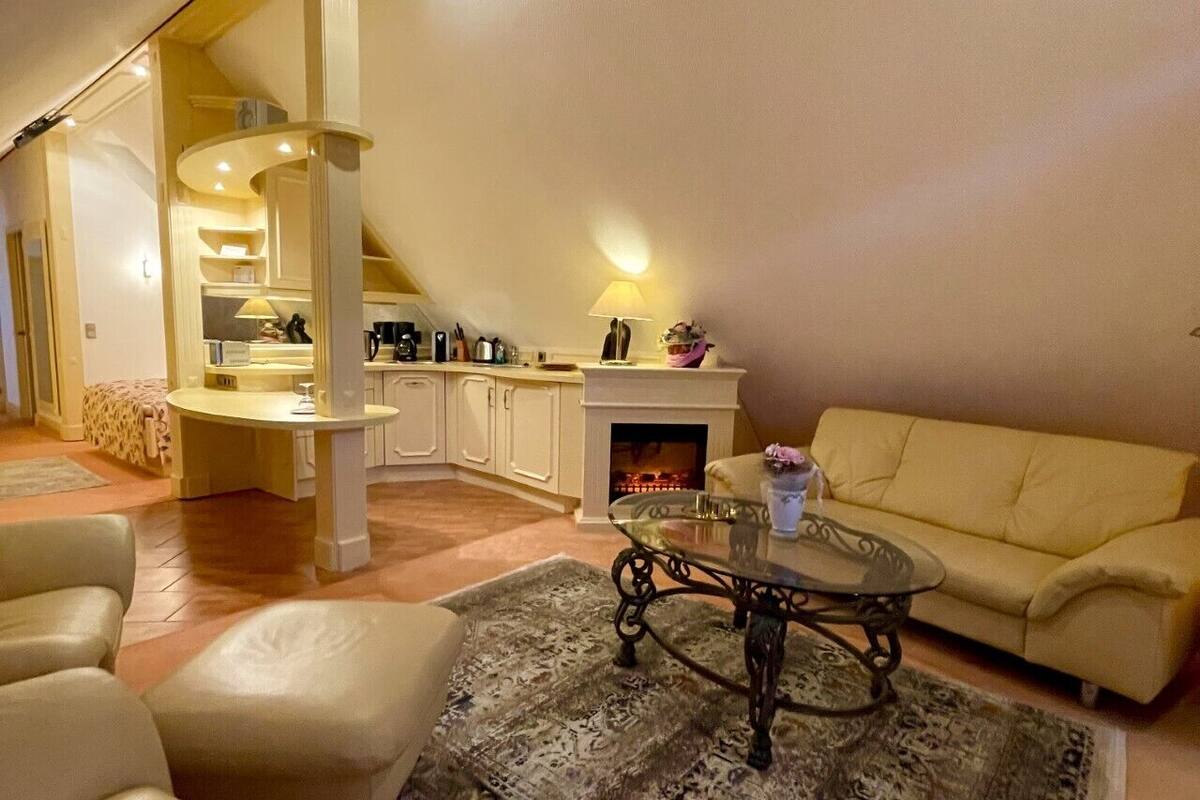A living room with a sofa, a coffee table, and a patterned rug. There is a fireplace and a kitchen area in the background.