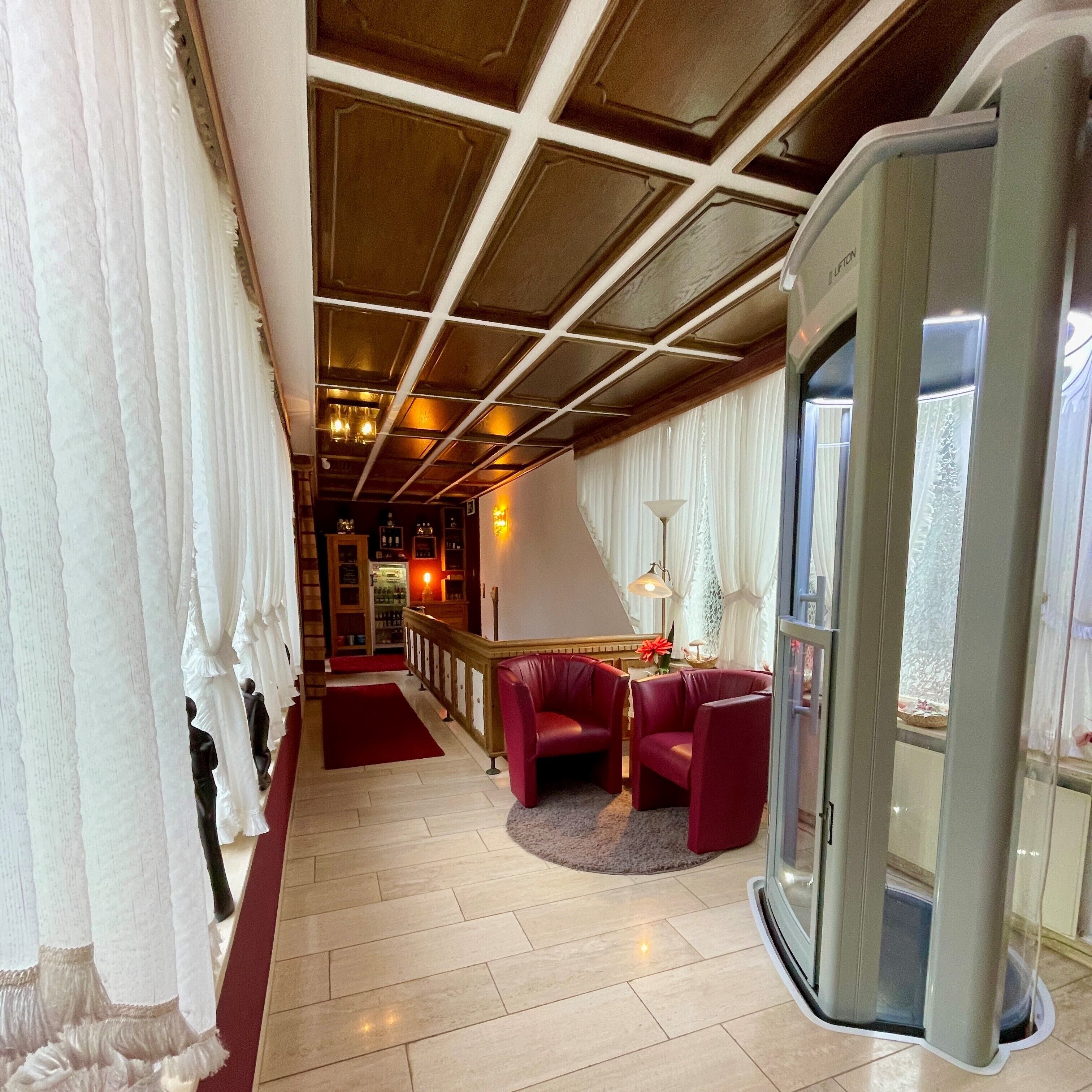 Un hall d’hôtel doté d’un plafond en bois, d’un comptoir de réception, d’un bar et d’un espace salon avec des fauteuils rouges.