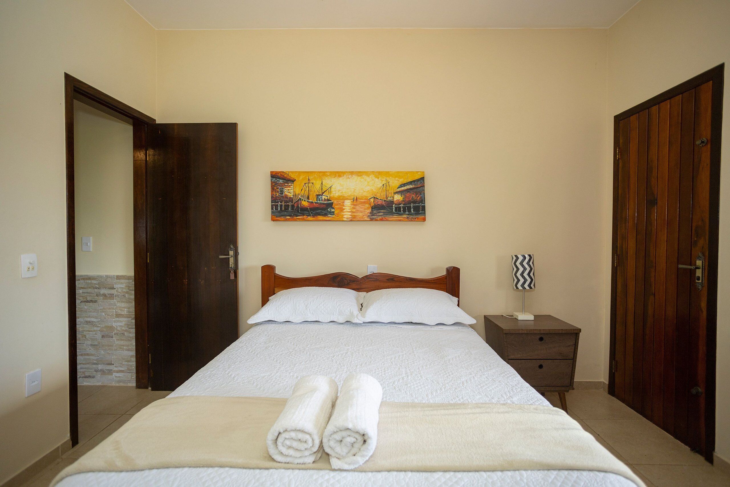 A neatly made bed with white linens and rolled towels, a wooden headboard, a painting of a harbor scene above the bed, and a nightstand with a lamp beside the bed.