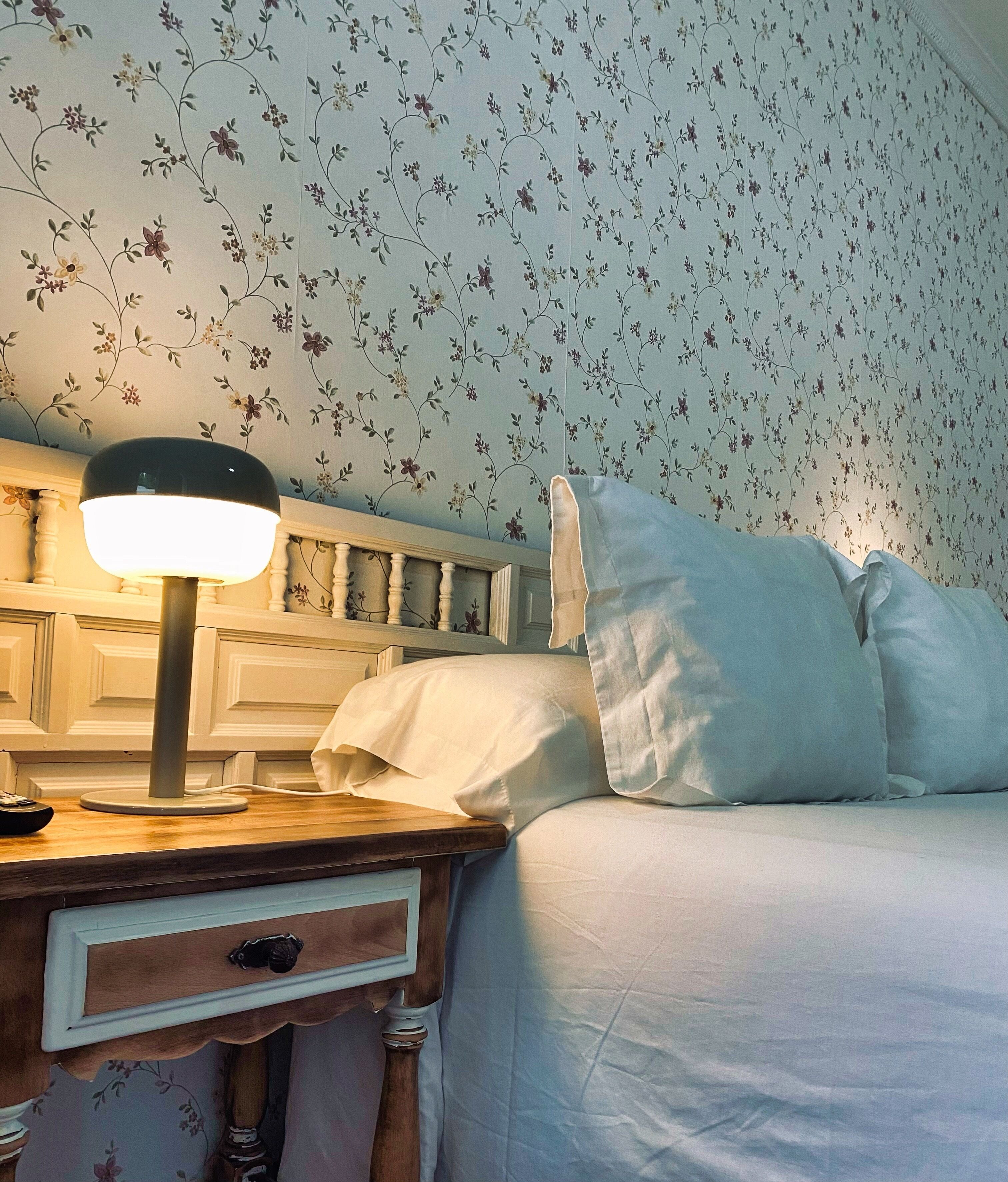 A neatly made bed with white linens, a bedside lamp, and a wooden nightstand with a drawer.