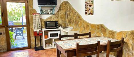 A dining area with a tiled floor, a table with chairs, a television on a shelf, and a stone wall.