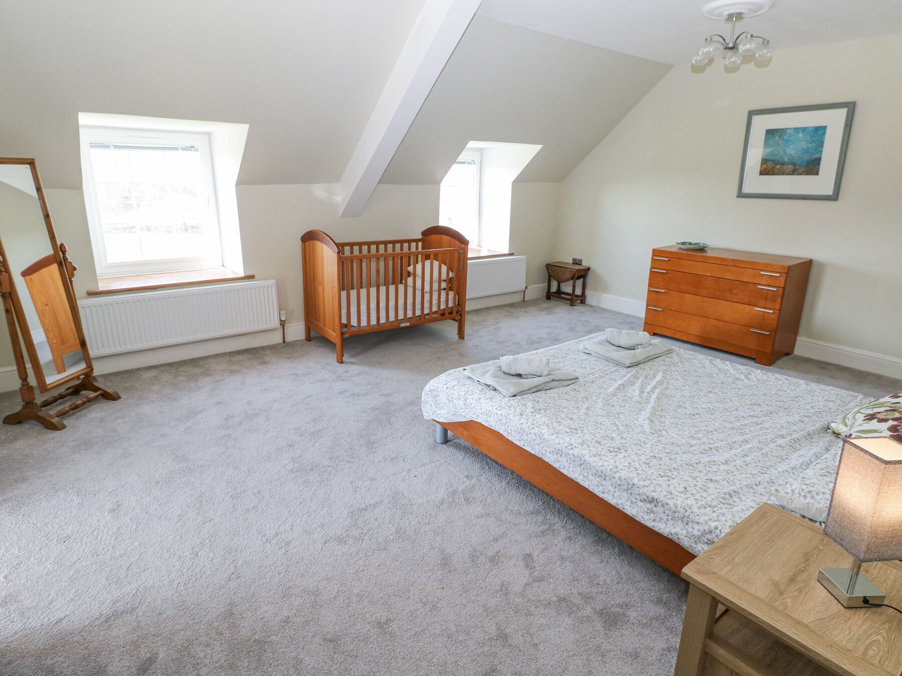 A bedroom with a sloped ceiling, a wooden crib, a bed with a grey bedspread, a wooden dresser, and a small table with a lamp.