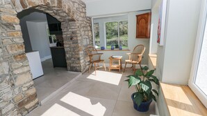 A stone archway leading to a kitchen, a dining area with chairs and a table, and a large window with a view of greenery. - Coytrahen House (Bridgend)