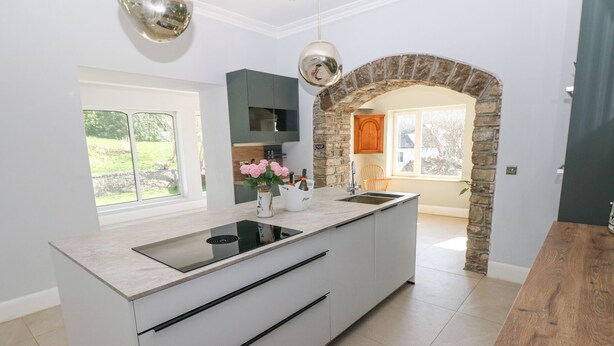 A modern kitchen with a central island, a stone archway, and a view of the outdoors. - Coytrahen House (Bridgend)