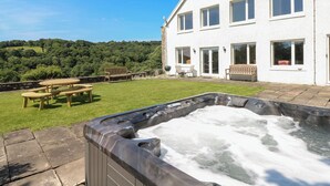 A house with a hot tub, a patio with a table and chairs, and a view of a green hillside. - Coytrahen House (Bridgend)