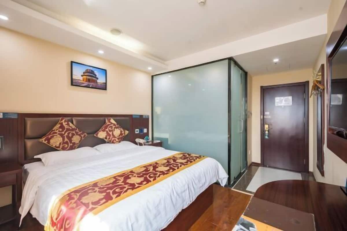 Una habitación de hotel con una cama, una mesita de noche, un separador de vidrio, una puerta y un cuadro en la pared.