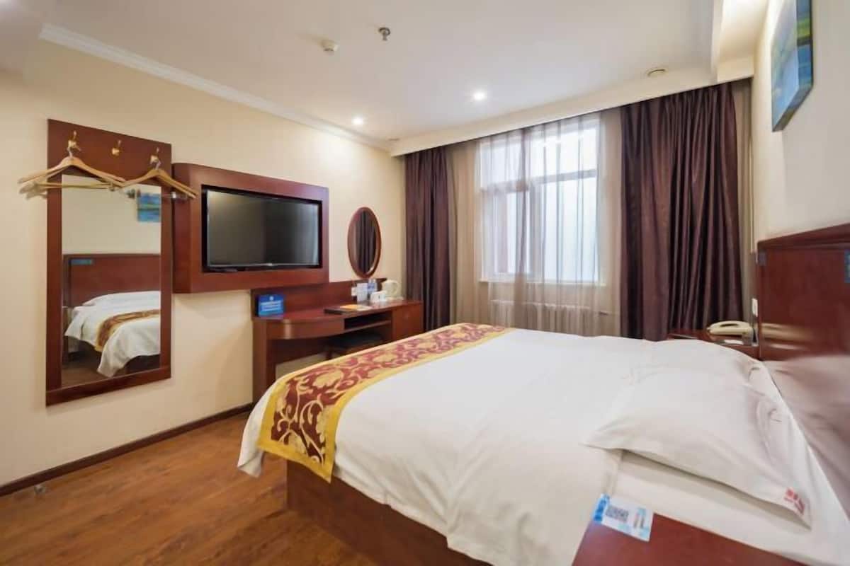 Habitación de hotel con una cama grande, un televisor montado en la pared, un escritorio con una silla, un espejo y una ventana con cortinas.