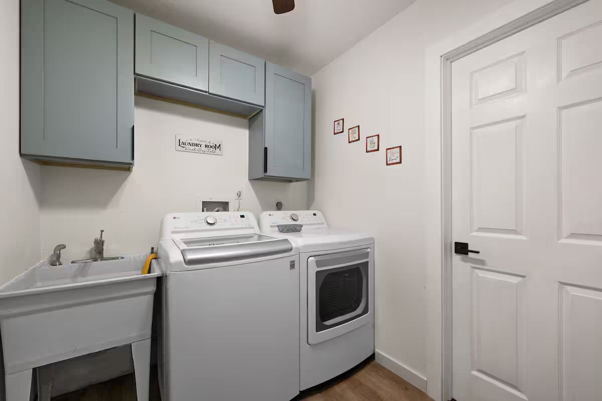 A laundry room with a washing machine, dryer, and a sink. There are light blue cabinets above the appliances and a white door to the right.