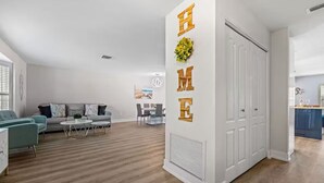 Ein modernes Wohnzimmer mit einer Couch, einem Couchtisch und Sesseln. Eine weiße Tür mit Lüftungsgitter und eine Wand mit der Aufschrift "HOME" sind zu sehen.