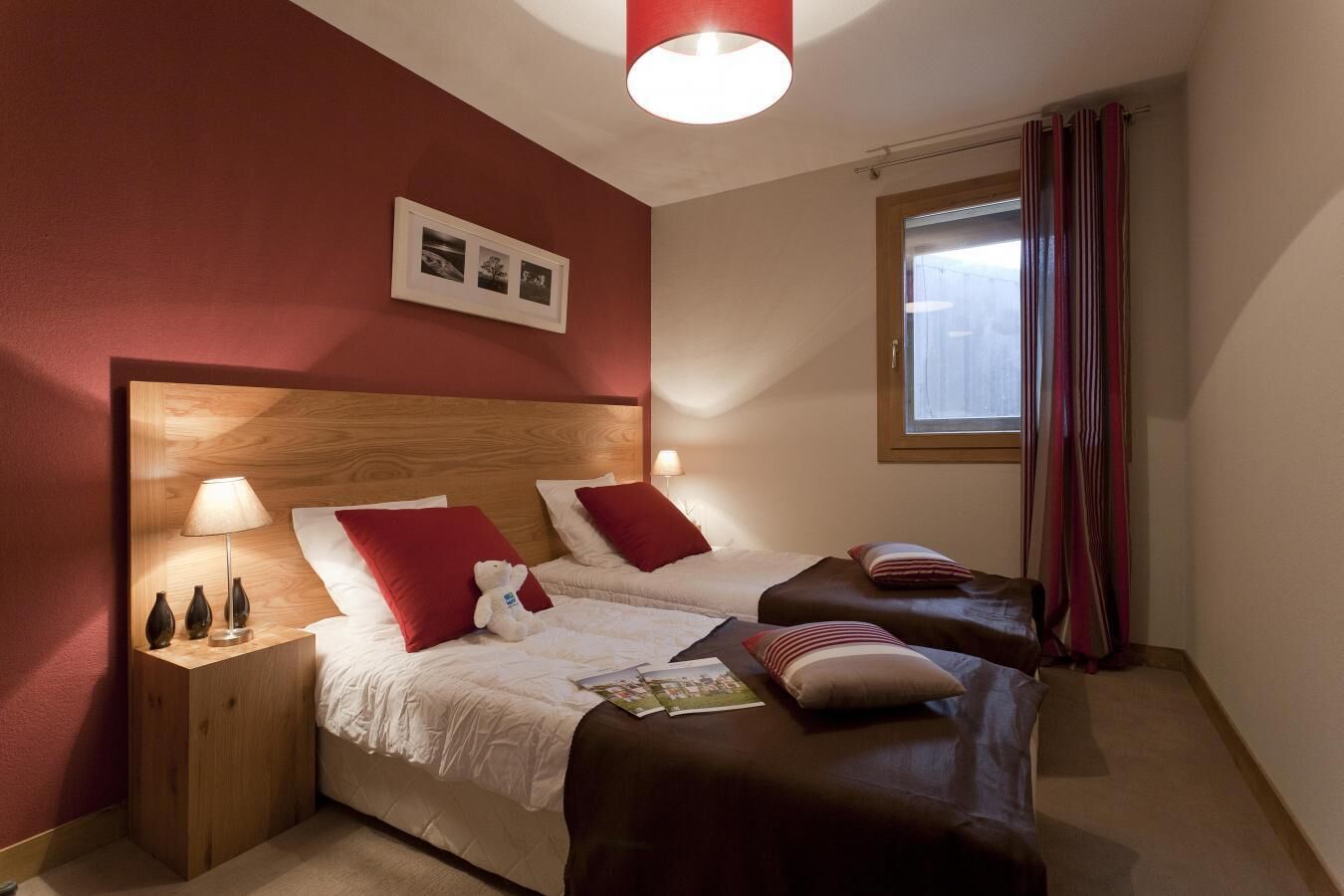A hotel room with two beds, a wooden headboard, a nightstand with a lamp, and a window with curtains.