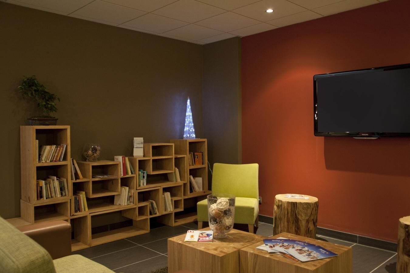 Et bibliotekshjørne med flere træbogreoler, en grøn stol, et rundt træbord med magasiner, og et ophængt fjernsyn.