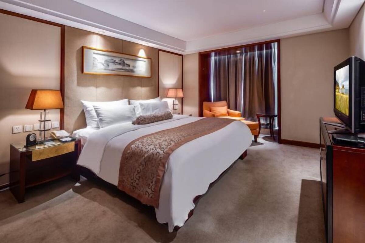 Ein Hotelzimmer mit einem großen Bett, einem Fernseher, einem Schreibtisch mit Lampe und einem Sessel.
