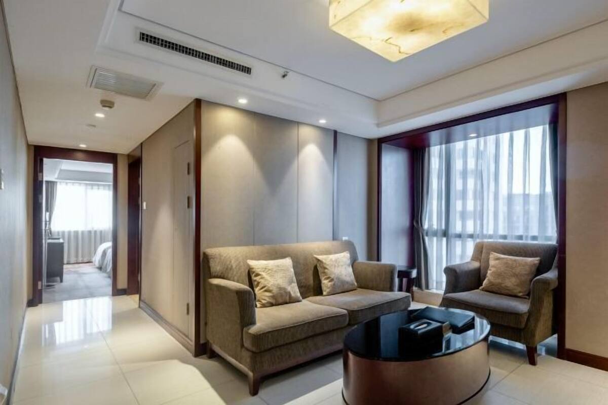 Ein modernes Wohnzimmer mit einer Couch, zwei Sesseln, einem Couchtisch und einem großen Fenster mit Vorhängen.
