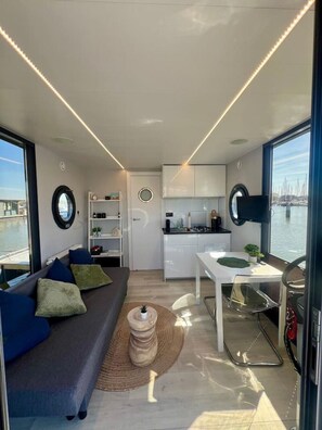 A compact interior with a sofa, dining table, kitchen area, and large windows offering a view of the water.