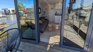 A modern interior with a glass door leading to a deck area. Inside, there is a sofa, a small round table, and a kitchen area. - Cosy Houseboat `Eclipse` in Marina Monnickendam (Monnickendam)