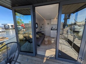 Un interior moderno con puerta de vidrio que da a una terraza. Adentro, hay un sofá, una mesita redonda y una zona de cocina.