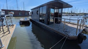 A modern boat house with a covered deck, a small window, and a dock extending into calm water. - Cosy Houseboat `Eclipse` in Marina Monnickendam (Monnickendam)