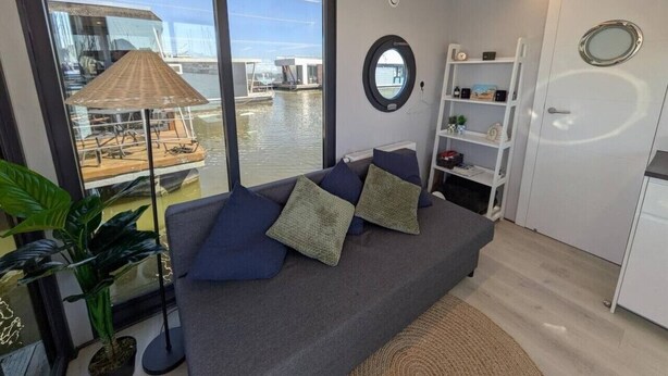 A modern living room with a sofa, a round mirror, and a view of a water body through large windows. - Cosy Houseboat `Eclipse` in Marina Monnickendam (Monnickendam)