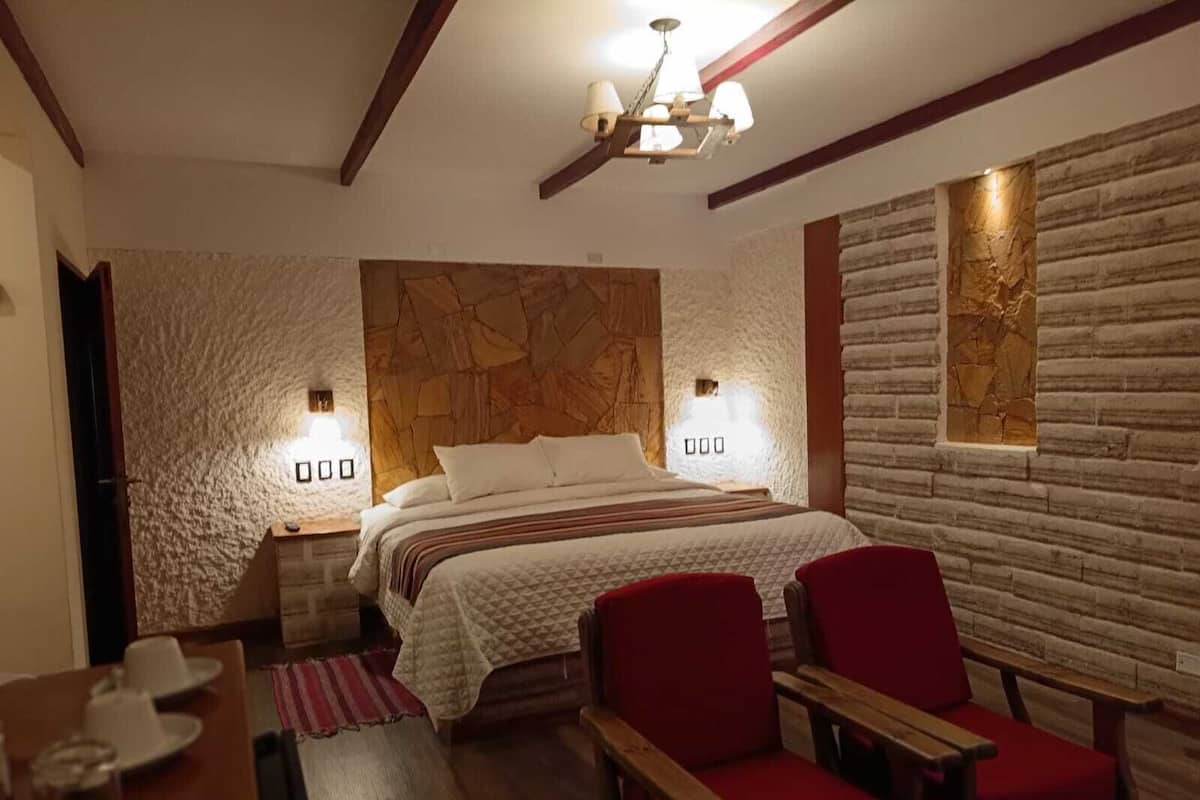 A bedroom with a large bed, two nightstands, a wooden headboard, a chandelier, and a stone wall.