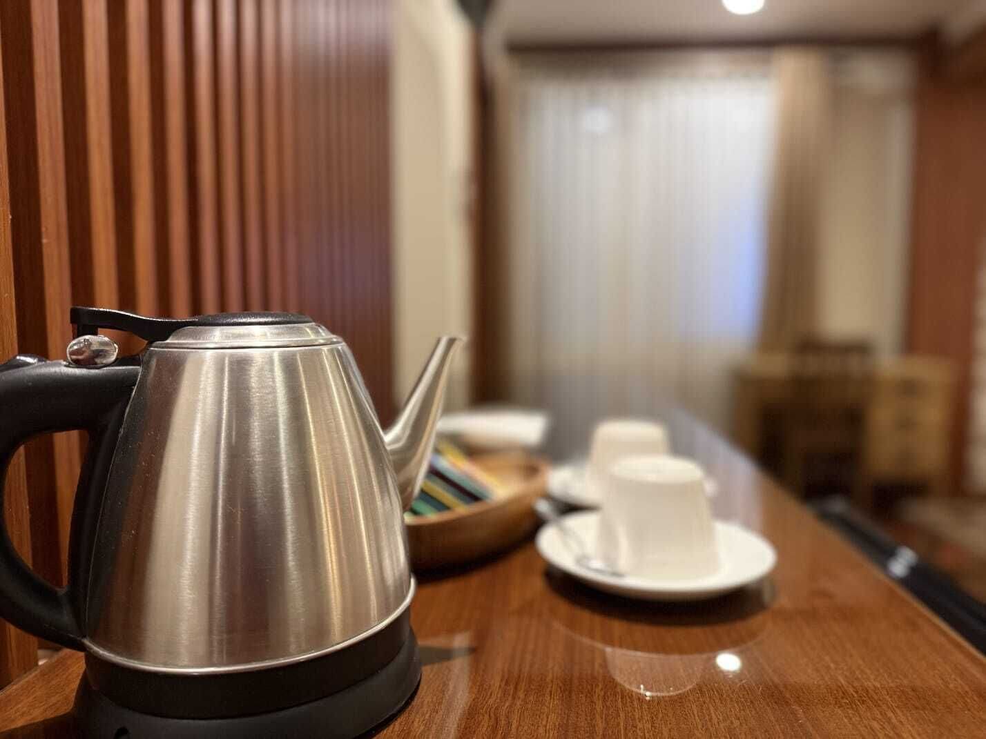 A stainless steel kettle on a wooden table with a cup and saucer in the background.