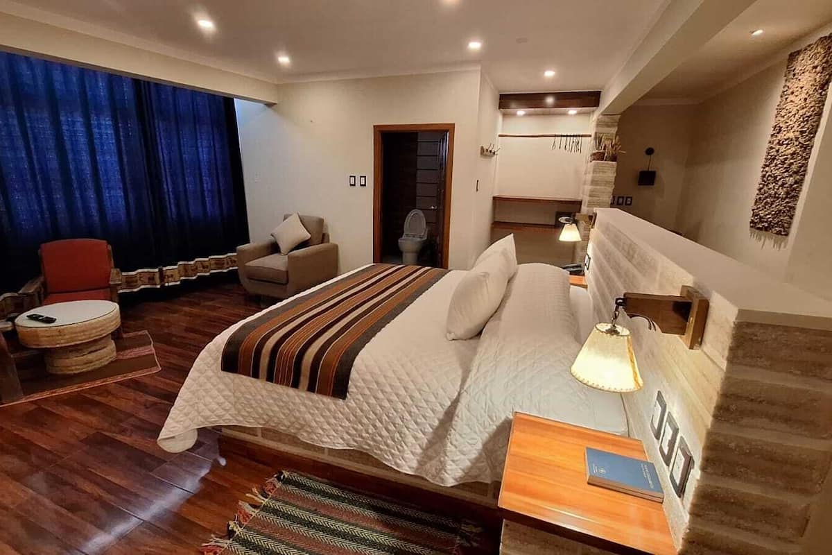 A spacious bedroom with a large bed, a sitting area with a chair and a small table, and a large window with curtains.