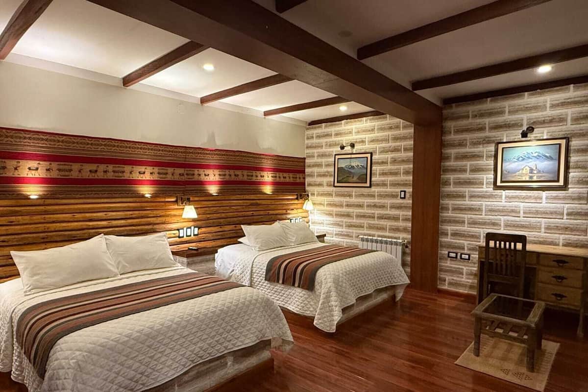 A hotel room with two beds, wooden headboards, a brick-patterned wall, a small table, and a chair.